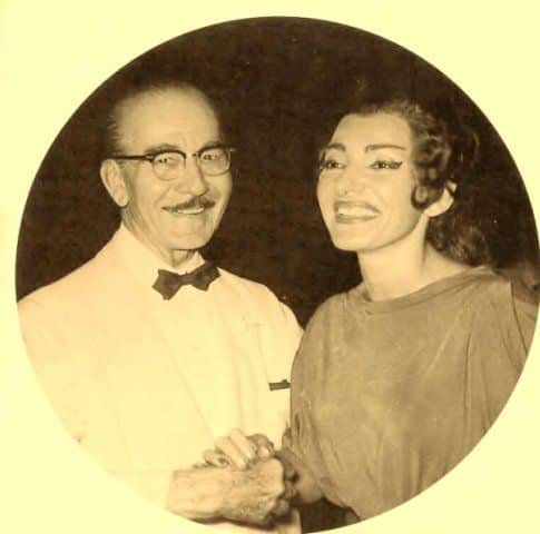 A sepia-toned photograph of Maria Callas smiling while holding hands with her father. He wears a light-colored suit and bow tie, while she is dressed in an elegant gown, both expressing joy and warmth.