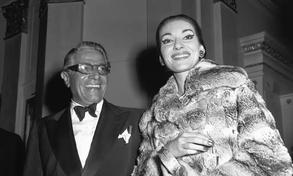Black-and-white photograph of Maria Callas standing next to Aristotle Onassis at a formal event. She is wearing a luxurious fur coat, smiling, while Onassis is dressed in a tuxedo, both looking glamorous and sophisticated.