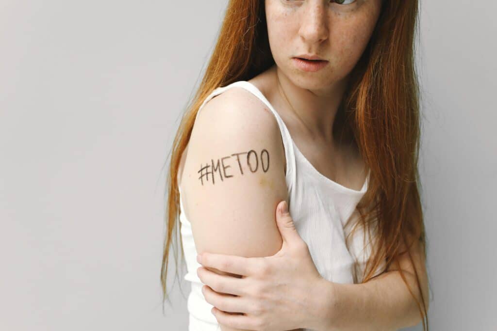 A young woman with long red hair looking off to the side, holding her arm where '#MeToo' is written. She wears a white tank top, and her expression conveys a sense of determination and vulnerability. The Trophy Of The Fight Is Justice.