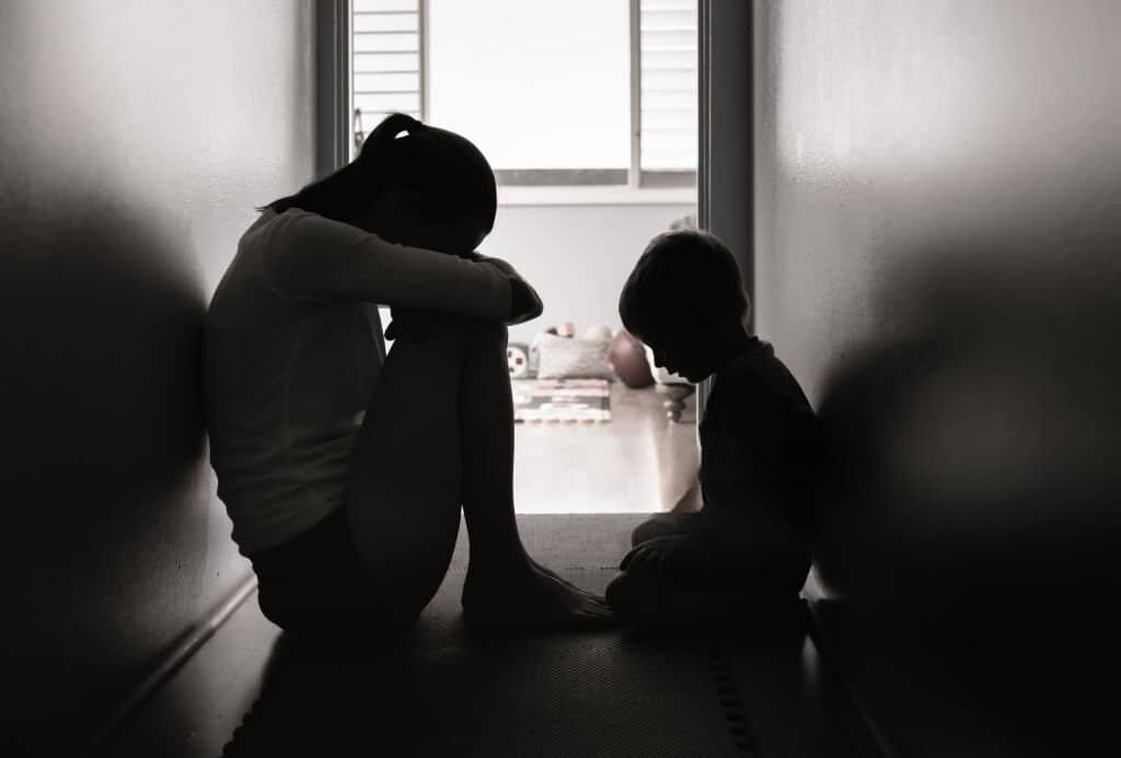 Silhouettes of a mother and child sitting in a hallway, both appearing withdrawn and sad.

