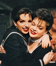 Color photo of Judy Garland embracing her daughter, Liza Minnelli. Both are smiling brightly and dressed in dark, stylish outfits. The image captures a moment of love and joy between the two, highlighting their close bond.