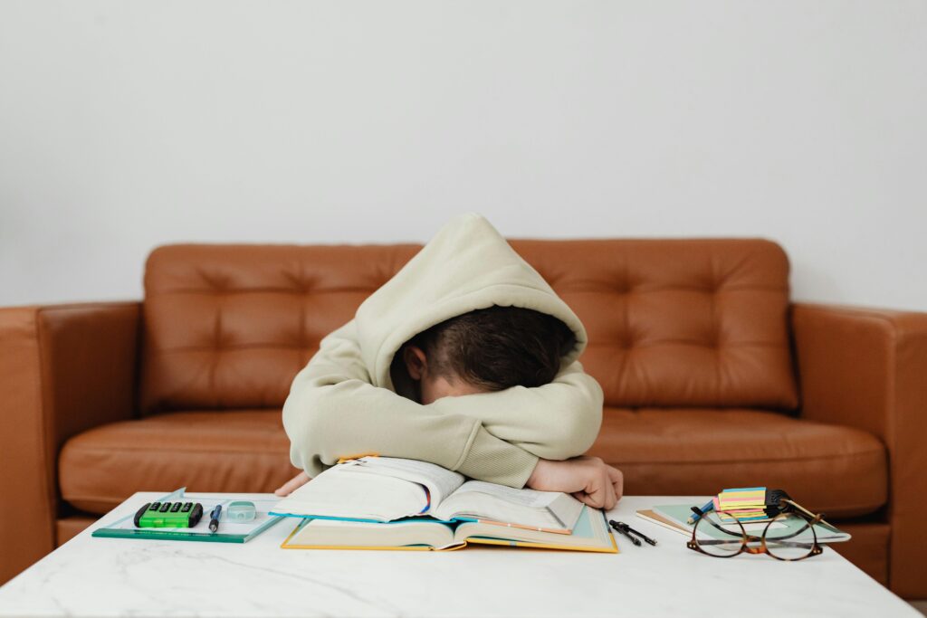A student wearing a hoodie resting their head on an open book, appearing exhausted from studying