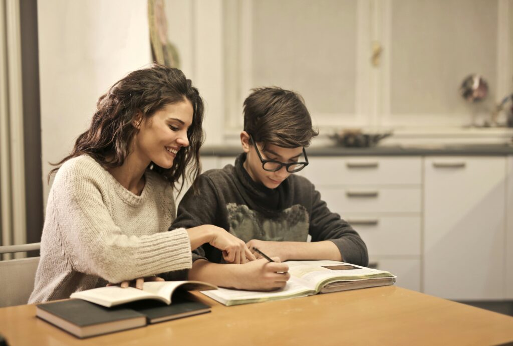 A woman smiling while helping a boy with his homework at a table.