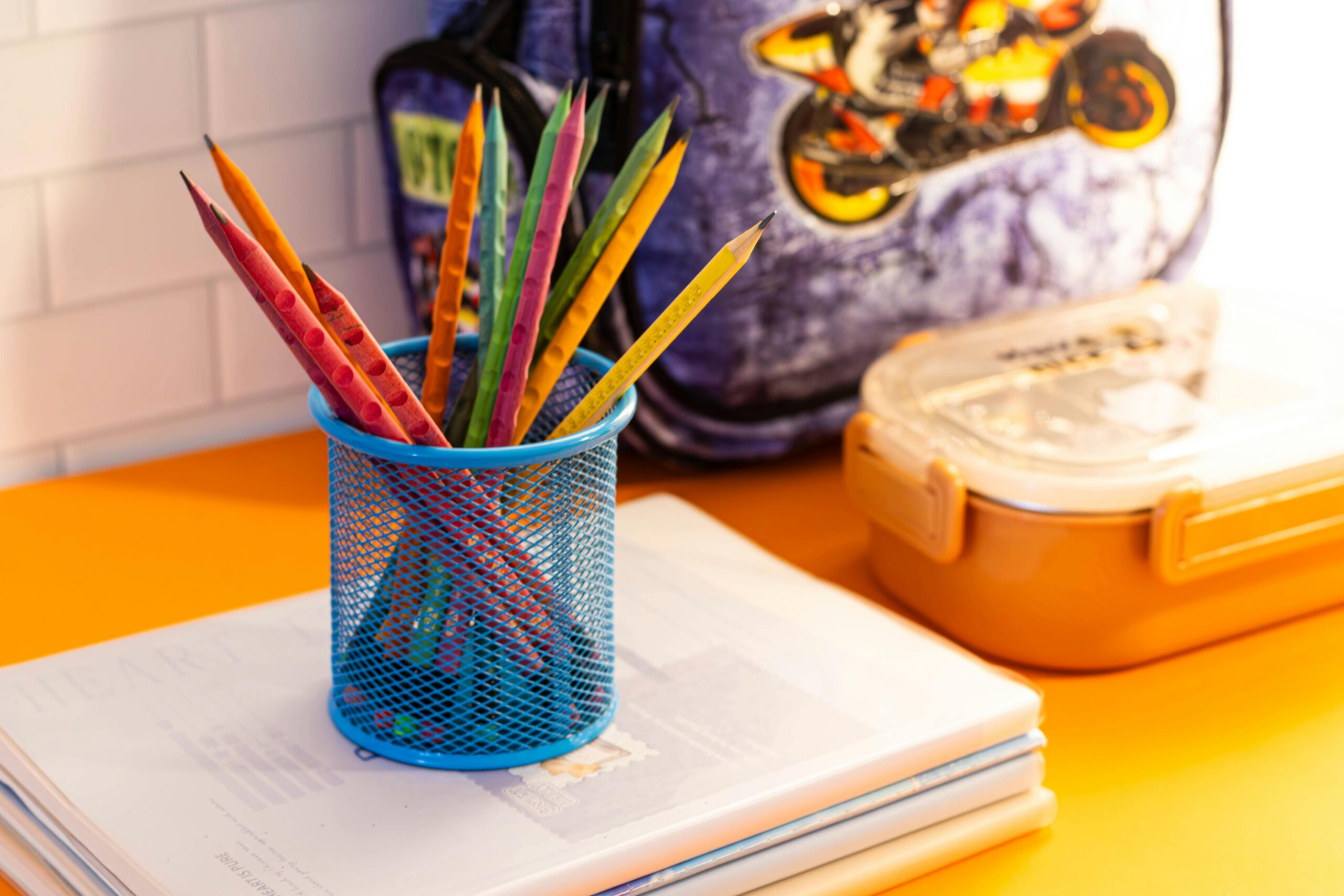 A colorful set of pencils in a mesh holder, notebooks, and a school backpack on a desk.